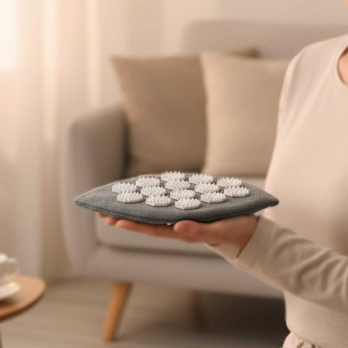 Person hält einen grauen Akupressur-Handschuh in Blattform mit weißen Massage-Spikes, im Hintergrund ein gemütliches Wohnzimmer.