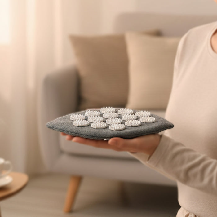 Person hält einen grauen Akupressur-Handschuh in Blattform mit weißen Massage-Spikes, im Hintergrund ein gemütliches Wohnzimmer.