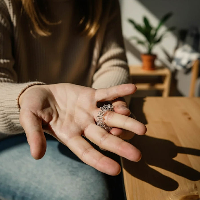 Person massiert mit einem silbernen Akupressur-Ring ihren Finger, sitzt am Holztisch im Sonnenlicht, im Hintergrund eine grüne Pflanze.