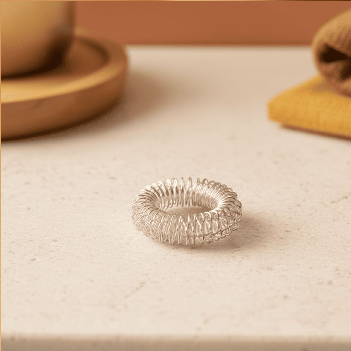 Silberner Akupressur-Ring aus Edelstahl liegt auf einer hellen Steinoberfläche, im Hintergrund unscharf eine Tasse und ein Tuch.