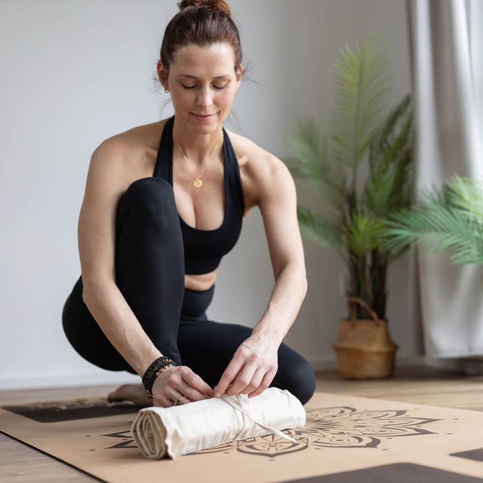 Frau in schwarzer Sportkleidung rollt beige Akupressurmatte auf gemustertem Yoga-Untergrund - Wellness-Übung im hellen, natürlichen Trainingsraum
