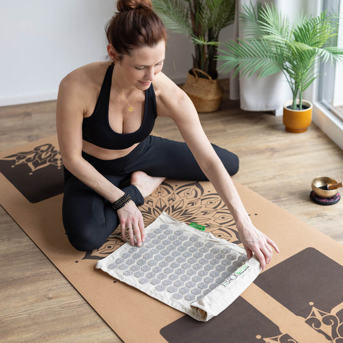Frau in schwarzer Sportkleidung bereitet Akupressurmatte auf Yoga-Unterlage vor - Meditationsraum mit Klangschale und grünen Pflanzen
