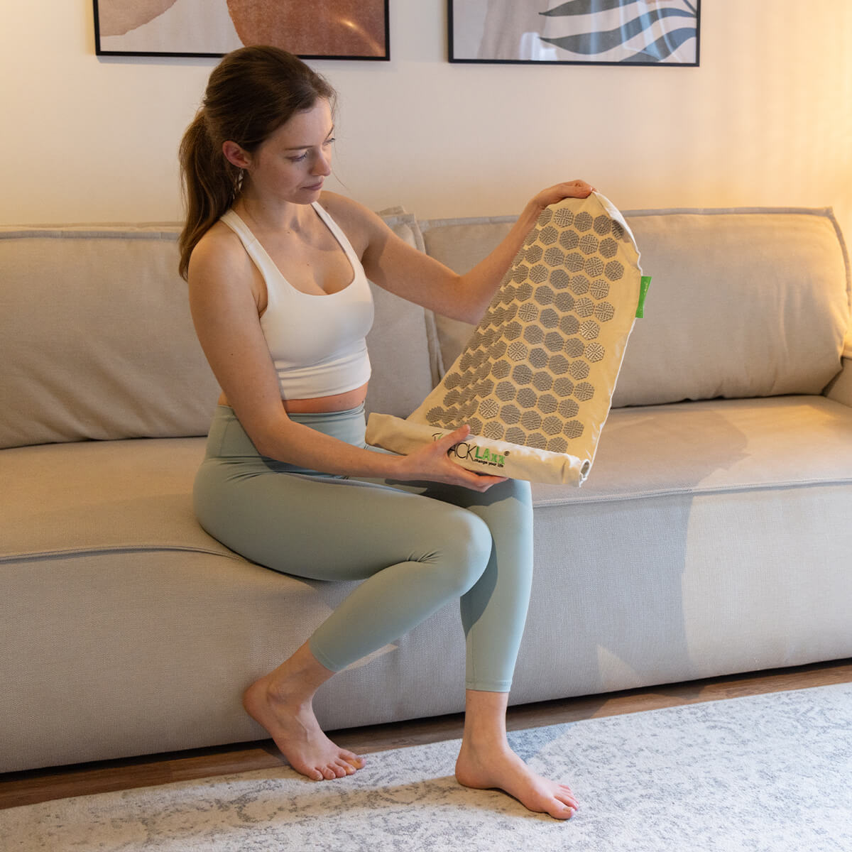 Frau im weißen Top hält Akupressurmatte mit hexagonalem Muster - Entspannungsprodukt für Wellness im gemütlichen Wohnzimmer auf beigem Sofa
