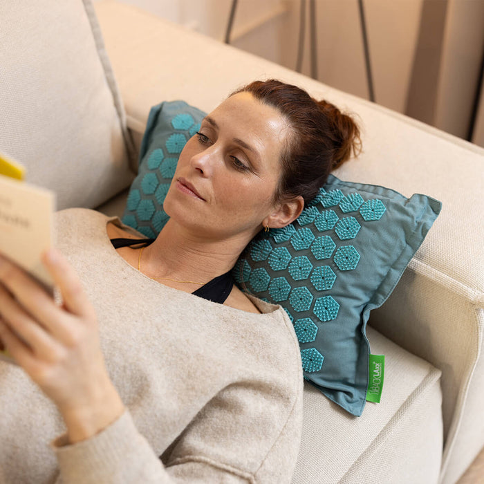 Person entspannt sich beim Lesen auf blauem BACKLAXX Akupressurkissen auf der Couch, natürliche Anwendung zuhause
