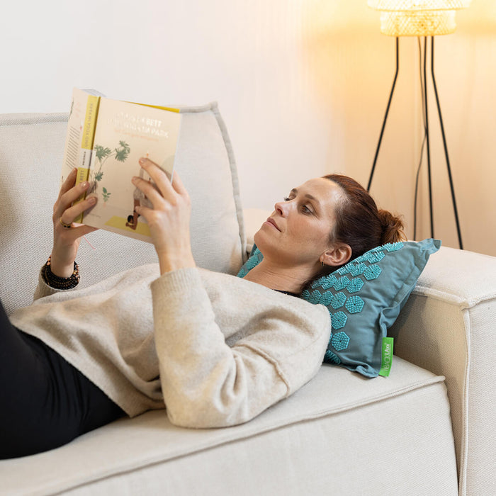 Person liest entspannt auf hellem Sofa mit türkisfarbenem ARTZT vitality Kissen - Gemütliche Lesestunde mit Stehlampe im Hintergrund
