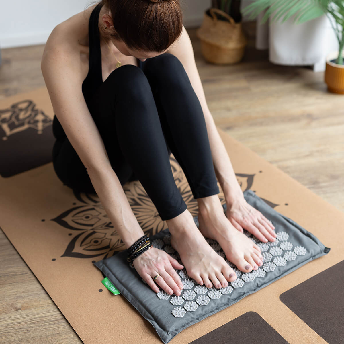 Frau sitzt entspannt am Boden, lehnt sich vor mit Armen um die Beine, Füße auf Akupressur-Fußmatte, schwarze Sportkleidung, meditative Yoga-Atmosphäre
