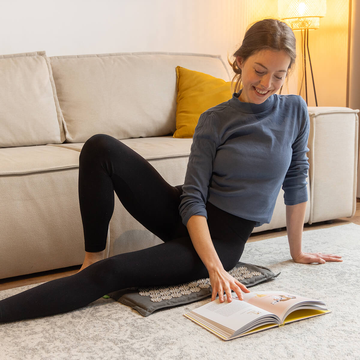 Frau sitzt am Boden im Wohnzimmer, liest Buch, steht auf Akupressur-Fußmatte mit runden Spitzen, blaues Langarmshirt und schwarze Leggings, Stehlampe im Hintergrund
