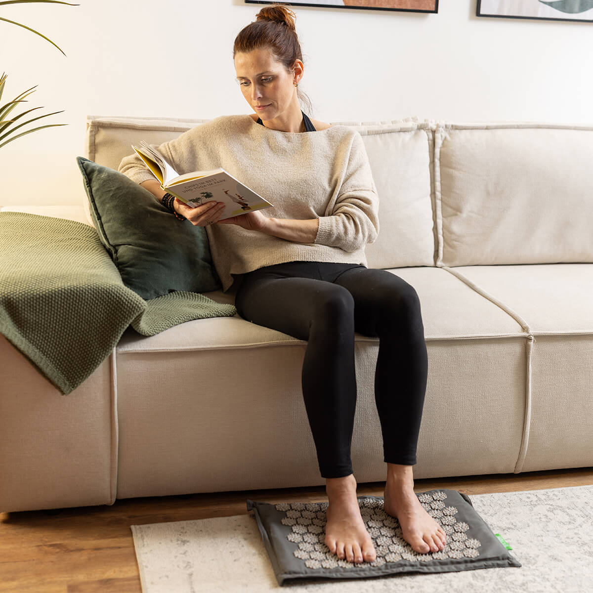 Frau sitzt entspannt auf beiger Couch und liest Buch, Füße stehen auf Akupressur-Fußmatte am Boden, trägt schwarze Leggings und beigen Pullover
