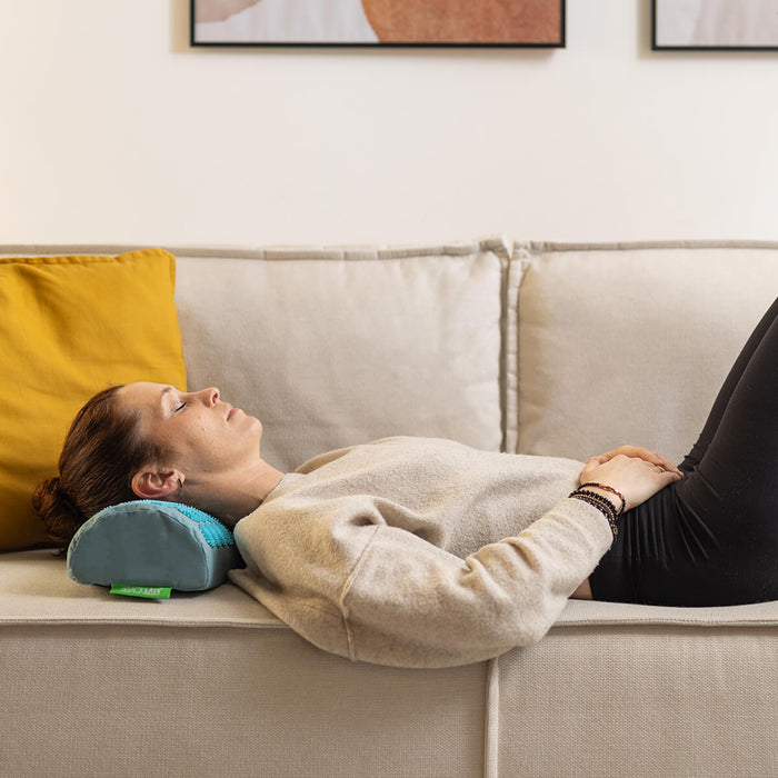 Person liegt auf Rücken auf Sofa mit blau-grüner Massage-Nackenrolle unter dem Nacken, beiger Pullover, entspannte Atmosphäre
