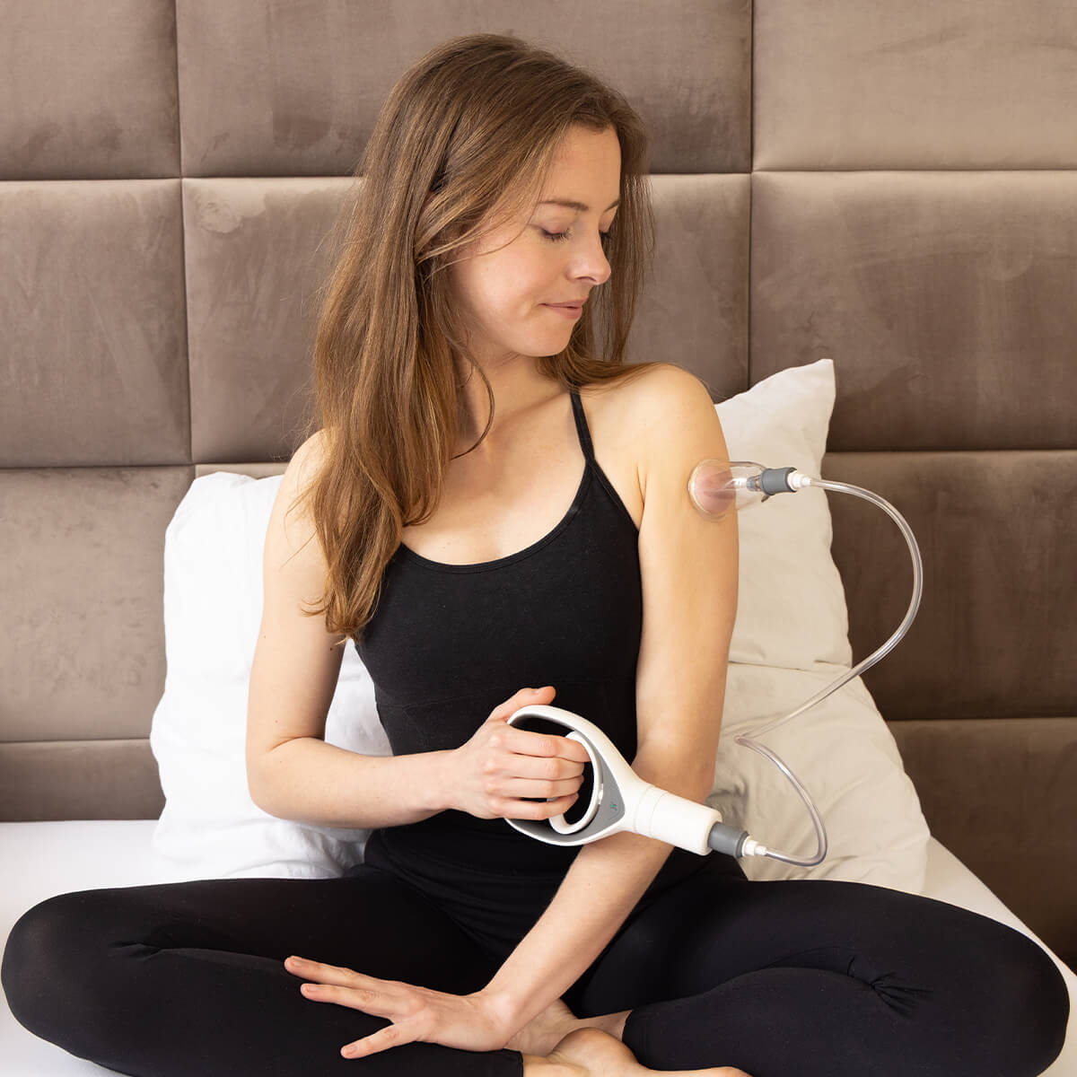 Selbst-Schröpfung am Arm: Person sitzt auf Bett und wendet Handpumpen-Gerät mit transparentem Schröpfglas am Oberarm an, schwarze Sportkleidung
