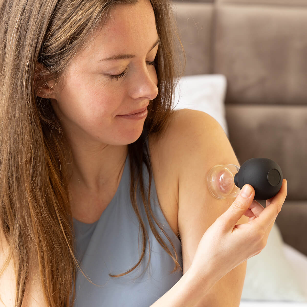 Frau in blauem Top hält BACKLAXX Schröpfglas-Set - Entspannende Anwendung von Vakuumtherapie im Schlafzimmer für Wohlbefinden und Muskelentspannung
