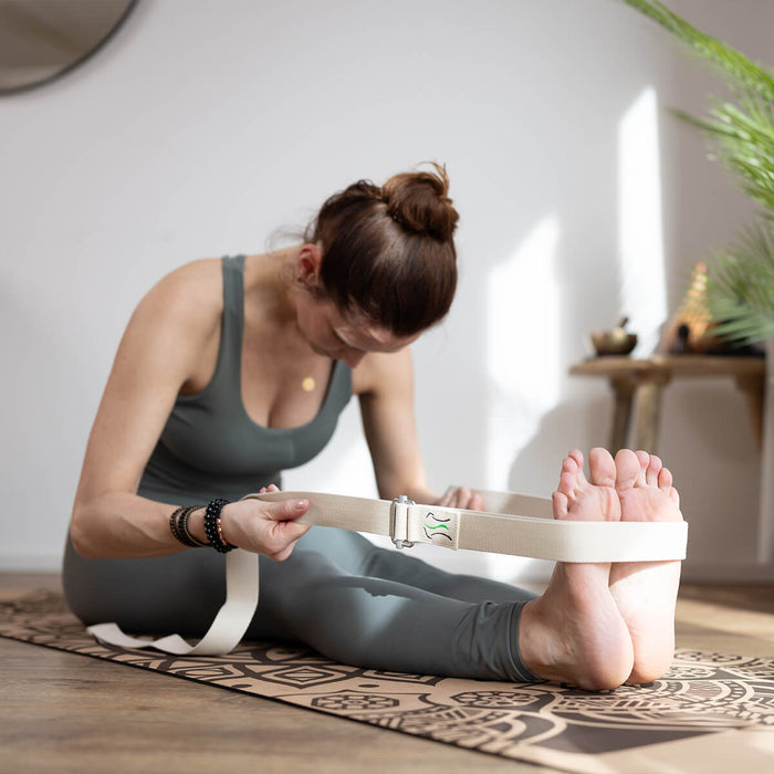 Frau sitzt auf gemusterter Yogamatte, macht Vorwärtsbeuge mit Yogagurt um die Füße, trägt graues Sport-Outfit, Pflanzen und Dekoration im Hintergrund
