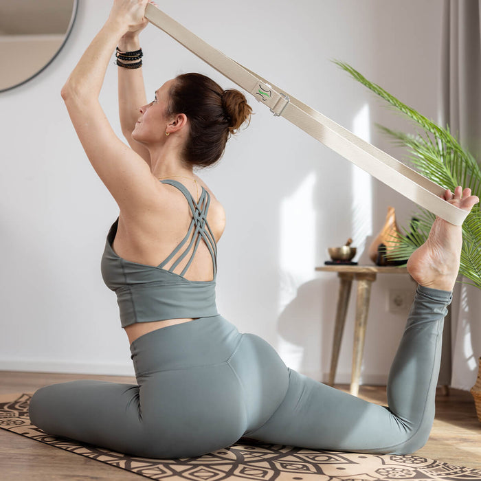 Frau sitzt auf Yogamatte, macht tiefe Dehnung mit Yogagurt, zieht Fuß zum Kopf, graue Sportkleidung, dezente Beleuchtung mit Dekoelementen
