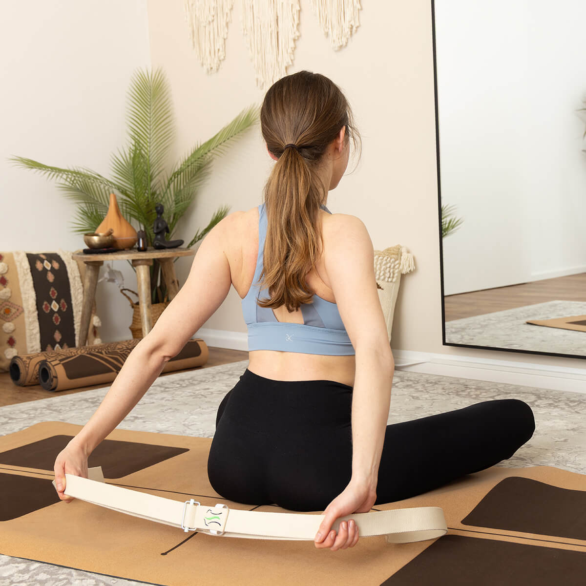 Frau sitzt im Schneidersitz auf Yogamatte, hält Yogagurt hinter dem Rücken, blaues Sport-BH, großer Spiegel und Wanddekoration im Hintergrund
