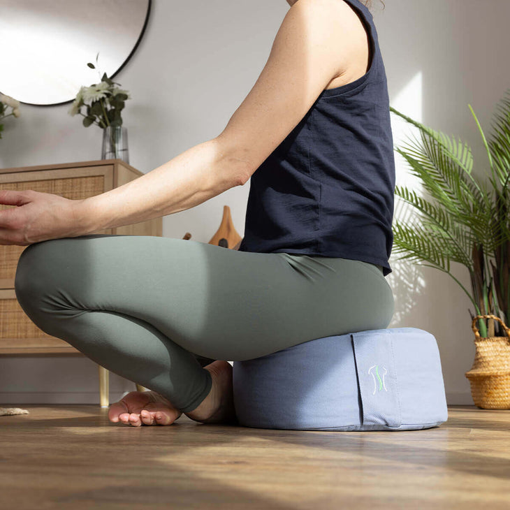 BACKLAXX Yogakissen in hellblau unterstützt meditative Sitzposition - Person praktiziert Entspannungsübung im gemütlichen Wohnraum mit Dekopflanzen
