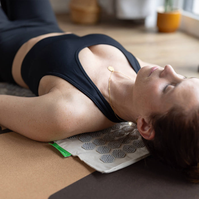 Frau in schwarzer Sportkleidung entspannt mit Kopf und Oberkörper auf beiger Akupressurmatte - Wellness und Entspannung im hellen Raum
