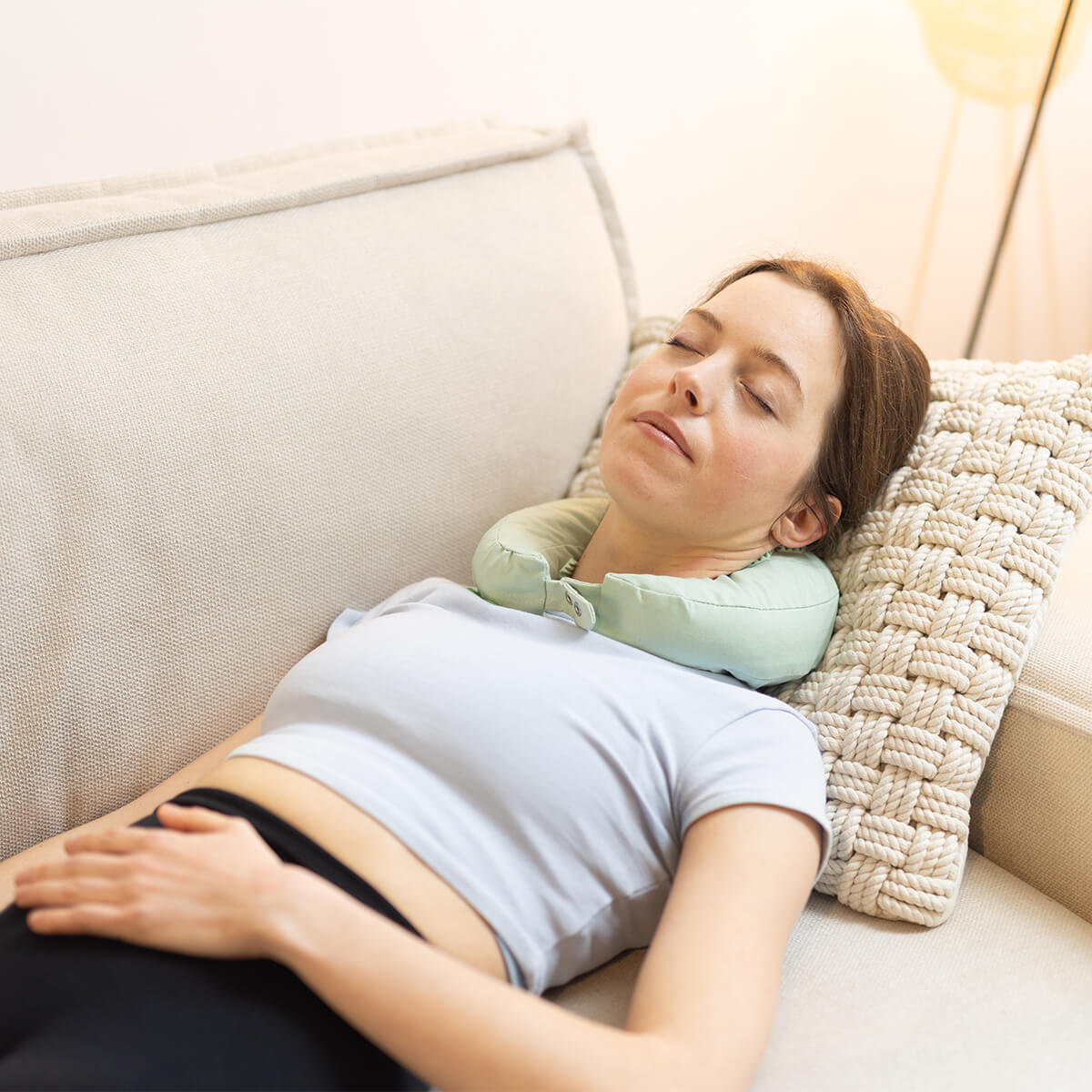 Person entspannt mit hellgrünem Nackenkissen auf beigem Sofa - Gemütliche Atmosphäre mit warmer Stehlampe für erholsame Ruhepause
